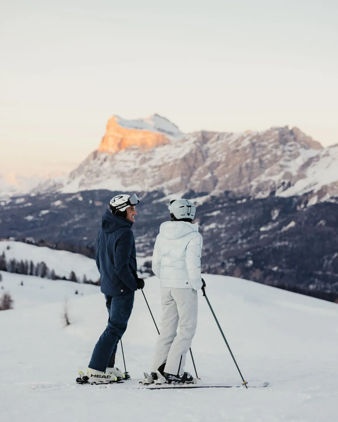 Two skiers in winter gear stand with poles on a snowy slope, mountains lit by sunset in the distance.