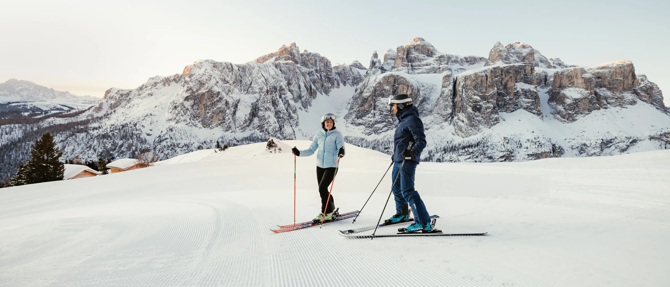 Due sciatori su una pista innevata conghia sulle Dolomiti al tramonto, montagne rocciose sullo sfondo.