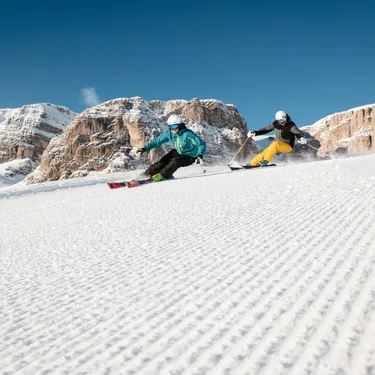 Due sciatori discendono una pista innevata tra molto ghiaccio, con montagne rocciose innevate sullo sfondo.