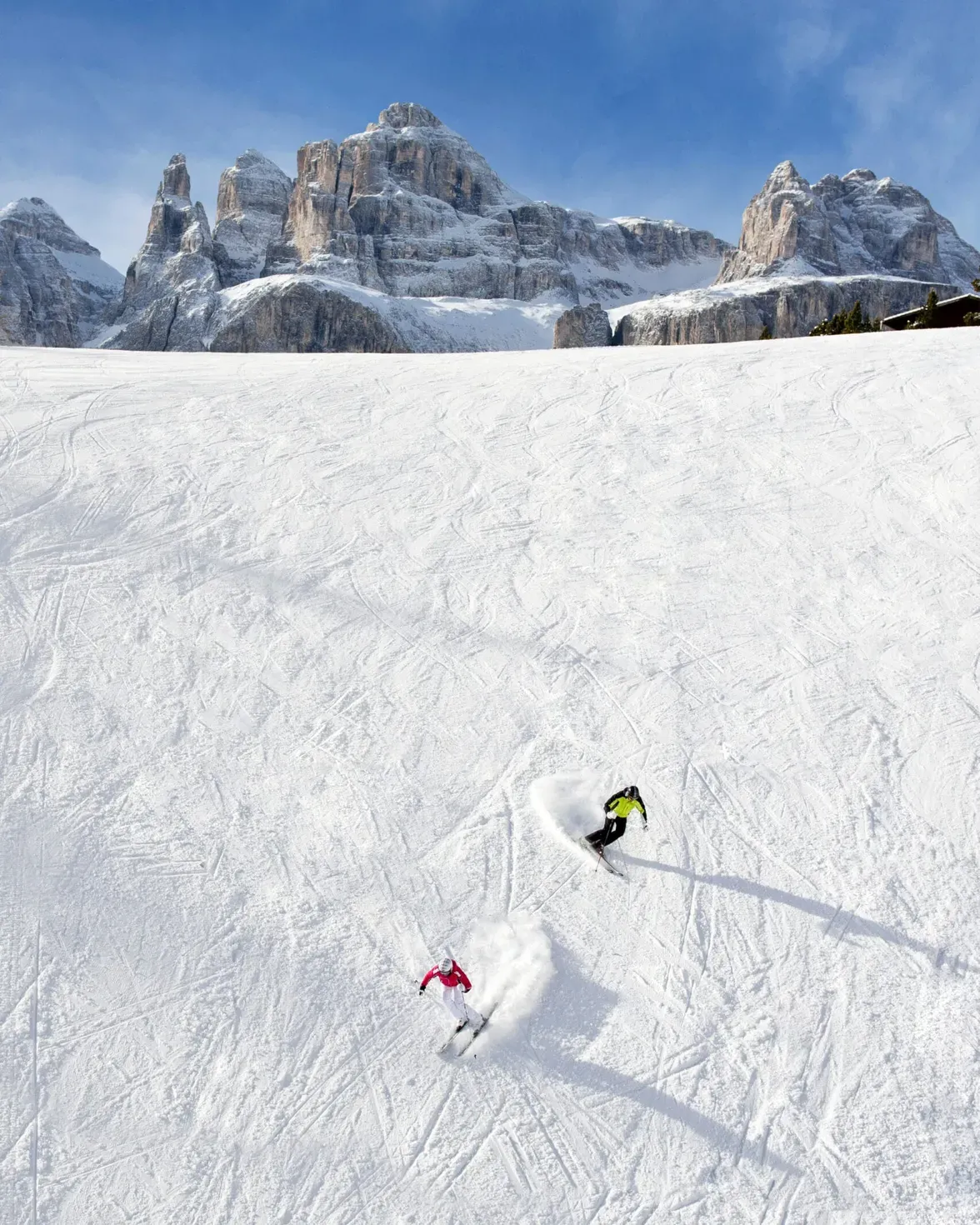 skiers carving down a snowy slope with rugged mountain peaks in the background under a blue sky