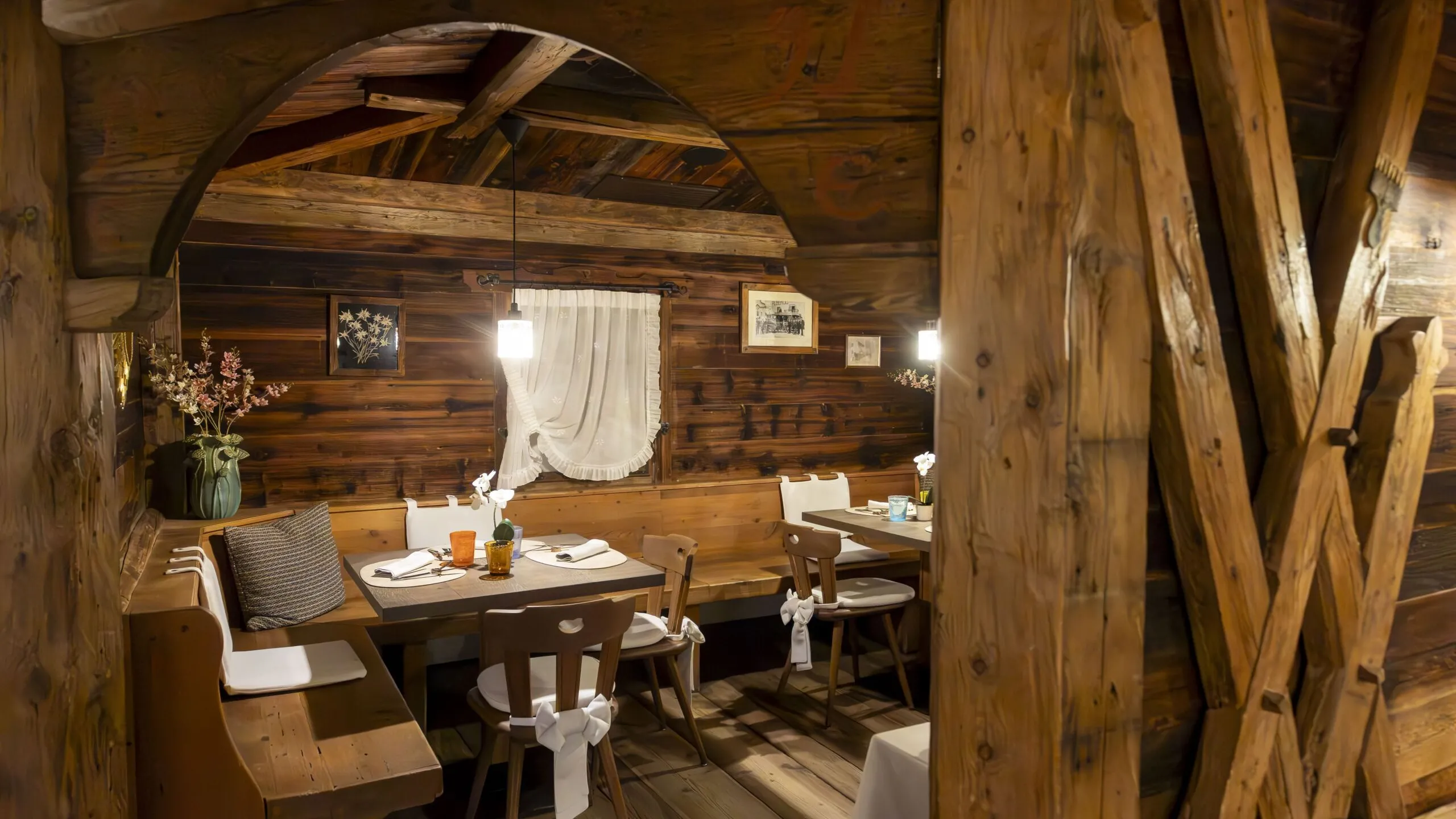Cozy rustic dining nook inside a wooden chalet with cushioned bench, tables set for guests, warm lighting, and decorative white ribbons on chairs.
