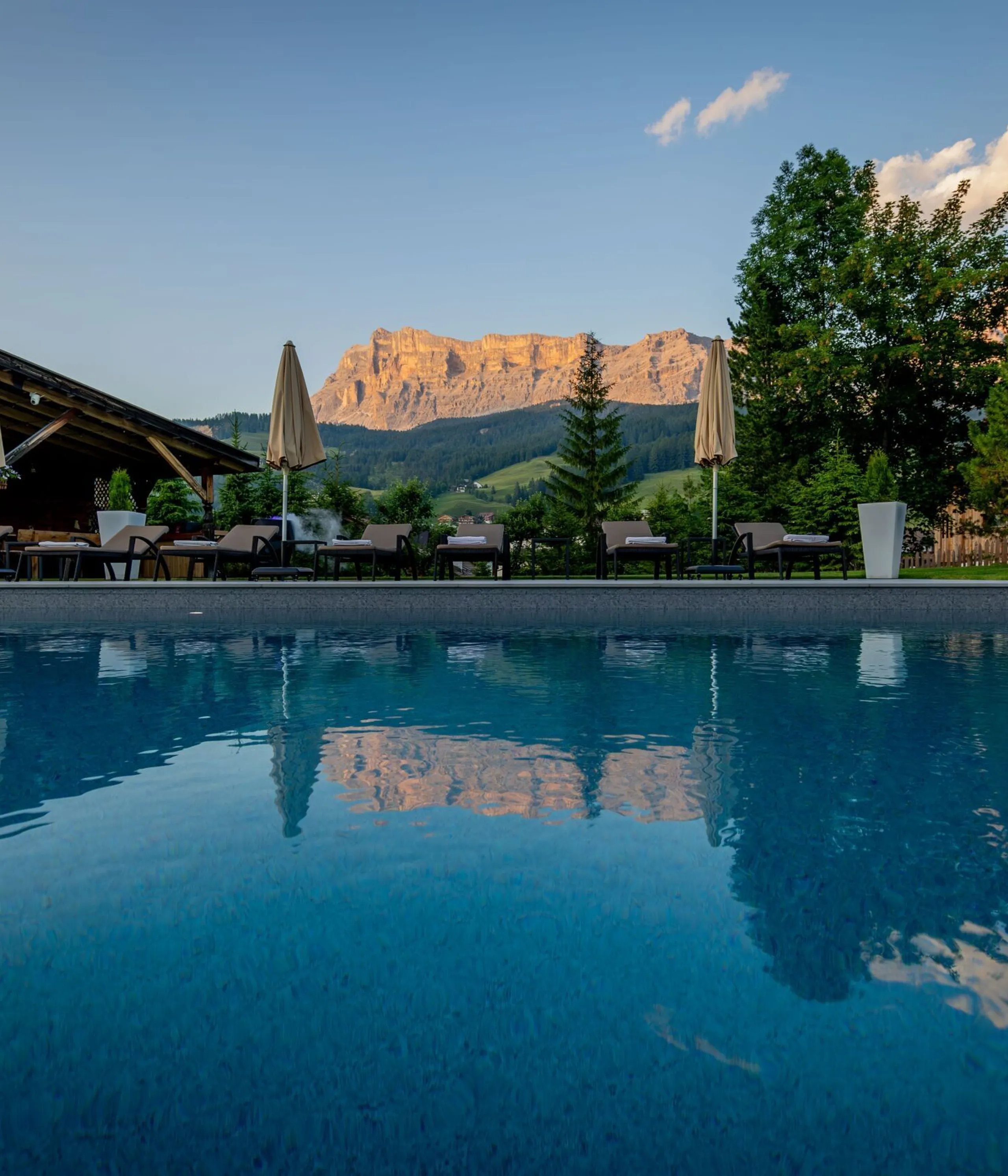 Serene outdoor pool with reflection of mountains, lounge chairs, and umbrellas against a wooden villa and green trees at dusk.